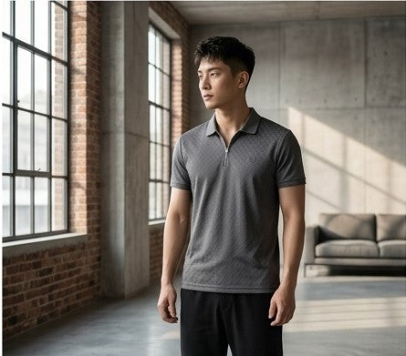 A man wearing a dark gray textured short-sleeve polo shirt with a half-zip collar, standing in a modern loft-style room with brick walls and large windows.