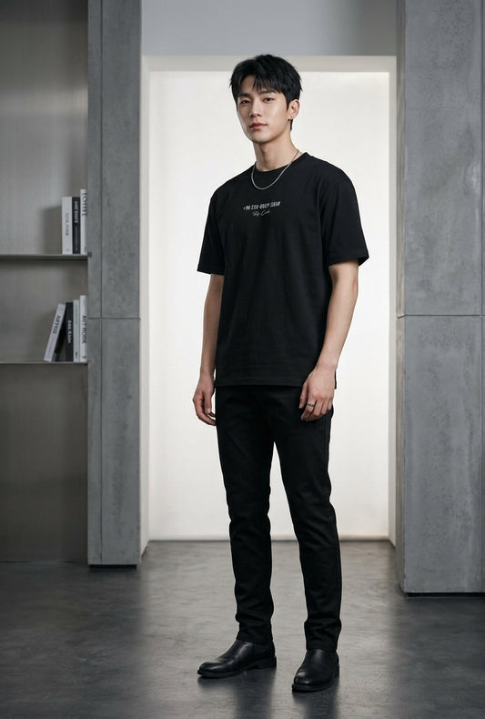A man wearing a black short-sleeve crewneck t-shirt with small white text on the chest, paired with black pants and black boots, standing in a modern gray interior setting.