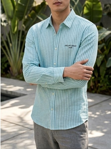 A man wearing a light blue and white vertically striped long-sleeve button-down shirt with 'MADAGASCAR' embroidered on the chest, standing outdoors with his arms crossed.