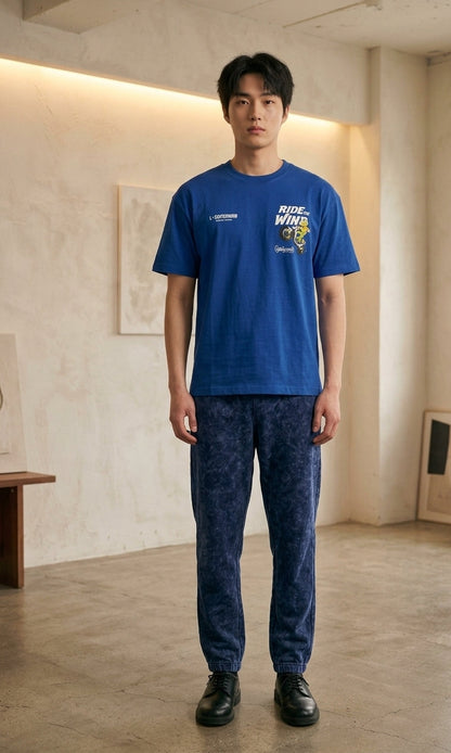 A man stands in a minimalist, warm-toned studio room wearing a royal blue graphic t-shirt and dark blue patterned trousers.