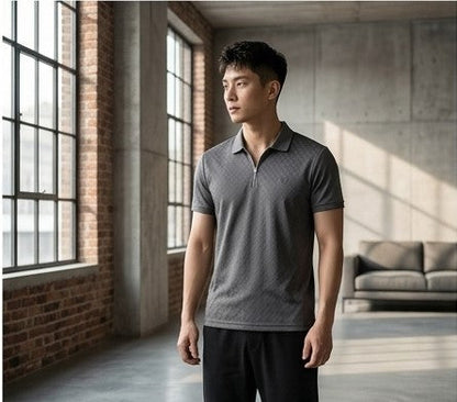 A man wearing a dark gray textured short-sleeve polo shirt with a half-zip collar, standing in a modern loft-style room with brick walls and large windows.