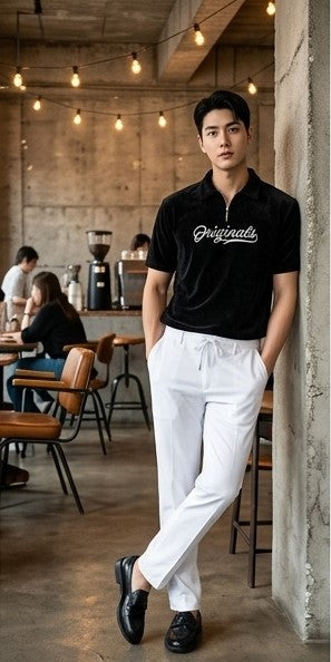 A man stands in a cafe wearing a black short-sleeve polo shirt with 'Grindslife' written in white script across the chest, paired with white drawstring trousers and black loafers.