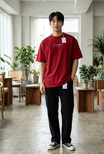 A man wearing a red crewneck t-shirt with white text and black pants, standing in a bright, modern indoor cafe or studio space with plants and wooden furniture.