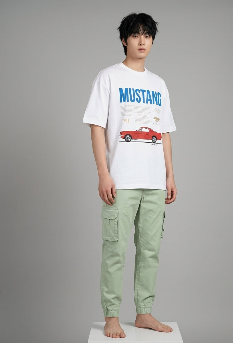 A male model stands barefoot on a white platform against a gray studio background, wearing a white graphic T-shirt with blue 'MUSTANG' text and a red car print, paired with light sage green cargo pants.