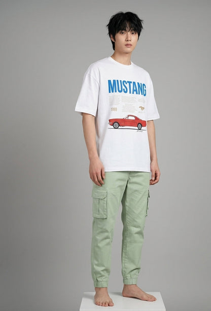 A male model stands barefoot on a white platform against a gray studio background, wearing a white graphic T-shirt with blue 'MUSTANG' text and a red car print, paired with light sage green cargo pants.