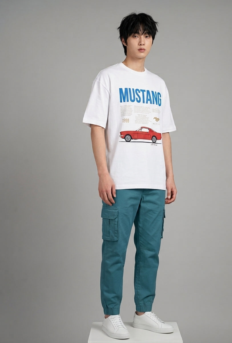 A male model stands against a neutral gray studio background wearing a white graphic t-shirt with a blue 'MUSTANG' text and red car print, paired with teal cargo pants and white sneakers.