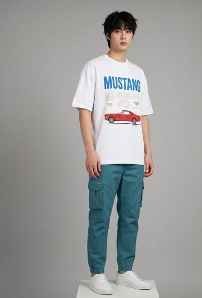 A male model stands against a neutral gray studio background wearing a white graphic t-shirt with a blue 'MUSTANG' text and red car print, paired with teal cargo pants and white sneakers.