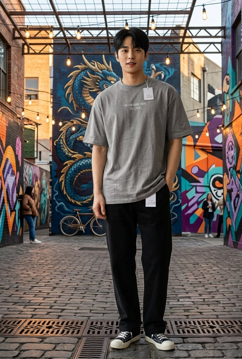 A man wearing a gray crewneck t-shirt with a small white graphic print, black pants, and black-and-white sneakers, standing in an outdoor alleyway with colorful graffiti murals on the walls.