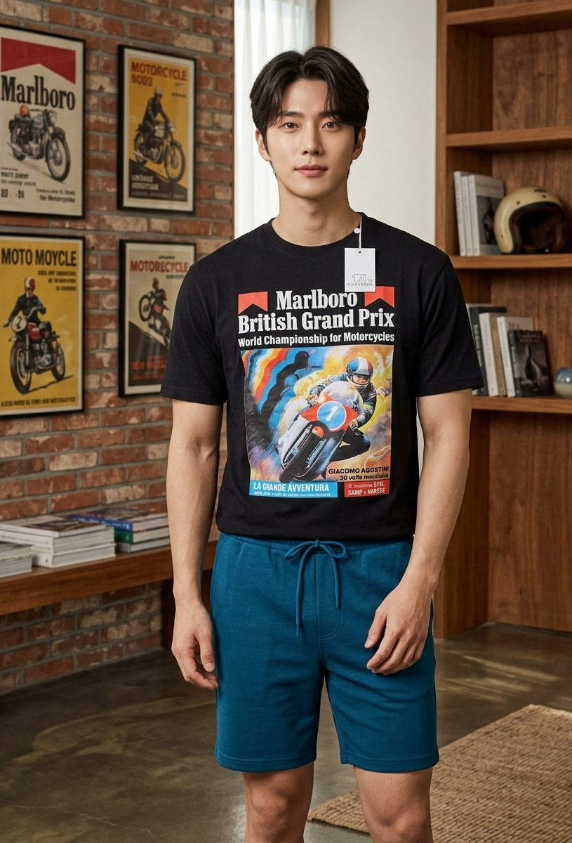 A man wearing a black t-shirt with a vintage-style Marlboro British Grand Prix graphic and teal drawstring shorts, standing in a rustic room with bookshelves and framed motorcycle posters.
