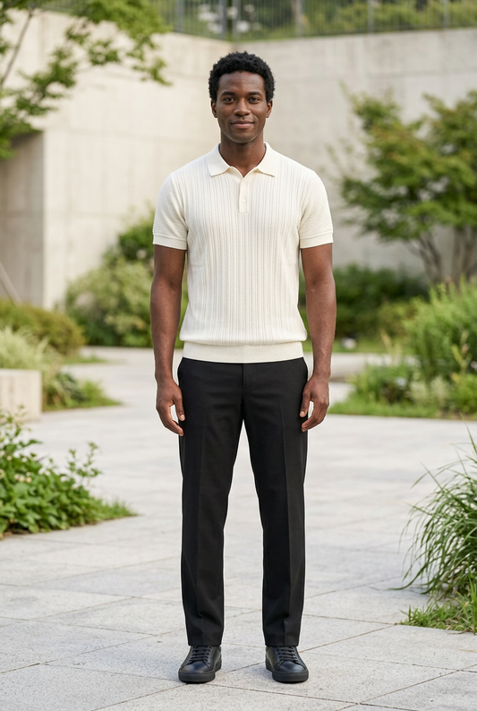 A man standing outdoors on a paved walkway wearing a cream ribbed short-sleeve polo shirt and black trousers.