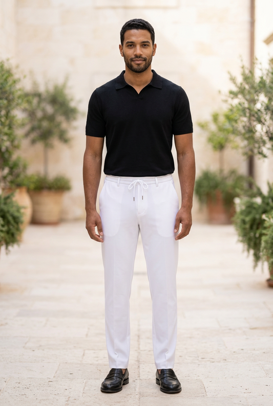 A man standing outdoors wearing a black short-sleeve polo shirt, white drawstring trousers, and black shoes, with a blurred stone wall and potted plants in the background.